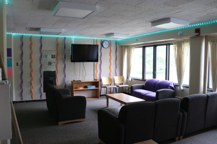 The image shows a cozy lounge area featuring a wall-mounted TV, a variety of seating options including armchairs and a small couch draped with a purple blanket, and a coffee table. The room is decorated with colorful streamers hanging from the walls and soft lighting around the ceiling.